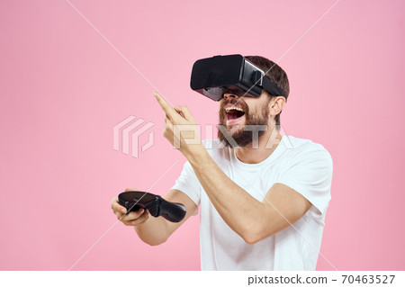 Man in virtual reality glasses joystick playing video lifestyle white t-shirt pink background Man in virtual reality glasses joystick playing video lifestyle white t-shirt pink background 70463527