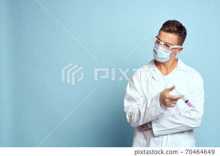 Doctor holds a syringe in his hand with a red liquid on a blue background medical gown and a protective mask Doctor holds a syringe in his hand with a red liquid on a blue background medical gown and a protective mask 70464649