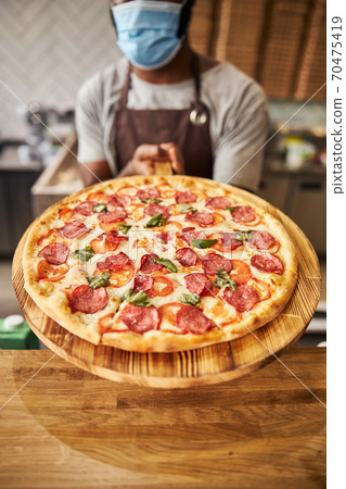 Male pizzeria worker in medical mask holding pepperoni pizza 70475419