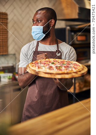 Afro American man in protective face mask holding pepperoni pizza Afro American man in protective face mask holding pepperoni pizza 70475421