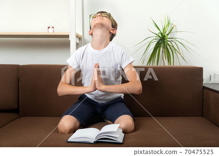 Boy on the couch prays to God next to book. Child is preparing for test exam. 70475525