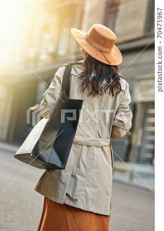 Rear view of a stylish woman wearing autumn grey coat and hat with big shopping bag walking city streets Rear view of a stylish woman wearing autumn grey coat and hat with big shopping bag walking city streets 70475967