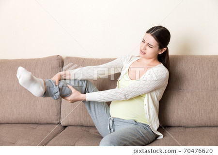 Leg cramps during pregnancy. Closeup of hands massaging swollen foot while sitting on sofa 70476620