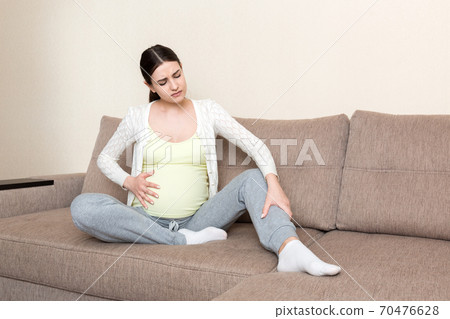 Leg cramps during pregnancy. Closeup of hands massaging swollen foot while sitting on sofa 70476628