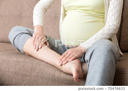 Leg cramps during pregnancy. Closeup of hands massaging swollen foot while sitting on sofa 70476633