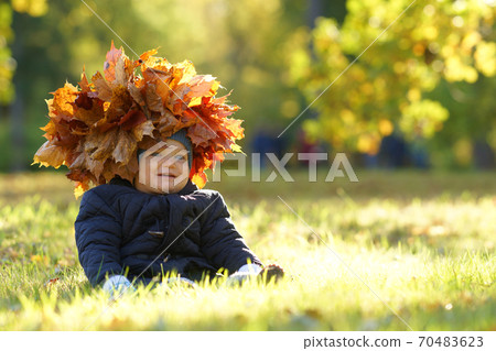 Little smiling child with wreath of leaves on head sitting on grass and laughing, background of sunny autumn park Little smiling child with wreath of leaves on head sitting on grass and laughing, background of sunny autumn park 70483623