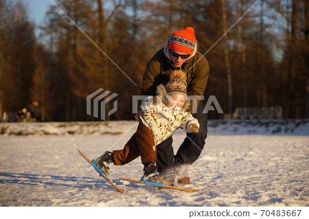 Young man learning his small child to ski. Smiling boy almast falling. Sunny photo on background of winter snowy park Young man learning his small child to ski. Smiling boy almast falling. Sunny photo on background of winter snowy park 70483667