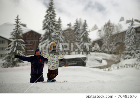 Small child on ski on mountain slope near smiling father and holding out hands. Background of snowy trees and houses Small child on ski on mountain slope near smiling father and holding out hands. Background of snowy trees and houses 70483670