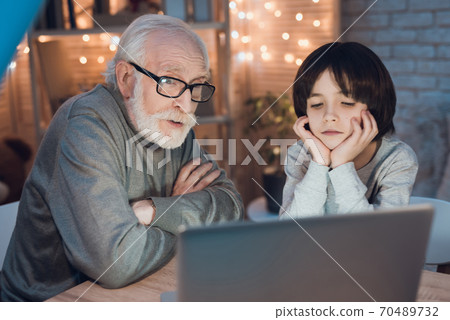 Grandfather and grandson are watching movie on laptop at night at home. Grandfather and grandson are watching movie on laptop at night at home. 70489732