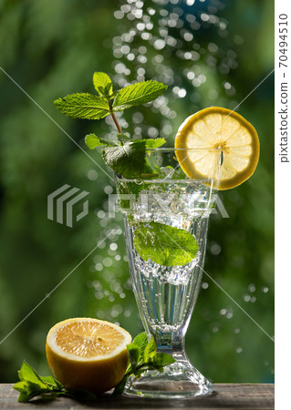 Sparkling water in a tall glass, ice cubes, mint and a slice of lemon, next to it is a lemon 70494510
