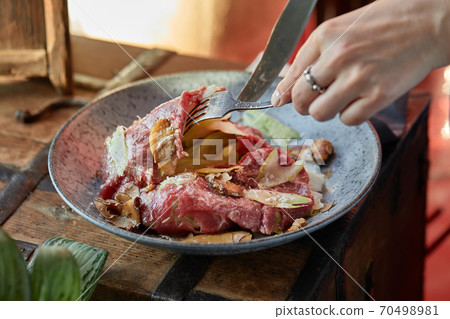 A female hand is preparing a vegetable salad with beef roast beef. A plate of salad with beef roast beef, onions, arugula, pepper and tomatoes on a black table. Top view. 70498981