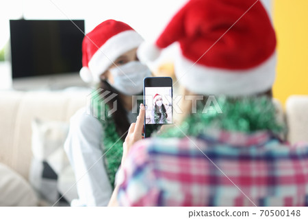 Woman photograph her friend on phone in santa claus hat and tinsel. Woman photograph her friend on phone in santa claus hat and tinsel. 70500148