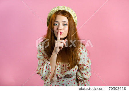 Smiling woman with red hair holding finger on her lips over pink background. Gesture of shhh, secret Smiling woman with red hair holding finger on her lips over pink background. Gesture of shhh, secret 70502638