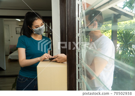 Asian man wearing a protective mask and delivering packages from grocery after online order. Woman accepting delivery boxes and Signing for the package in tablet. Concept of transportation service. 70502938