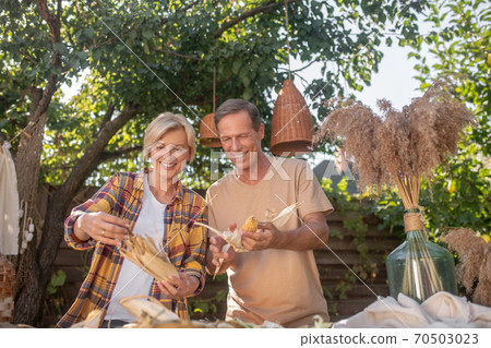 Happy middle-aged couple shucking corn outside, smiling 70503023