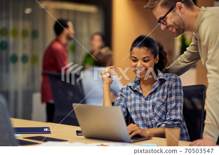 Young cheerful caucasian man helping female coworker with computer work, two young multiracial colleagues using laptop and discussing project Young cheerful caucasian man helping female coworker with computer work, two young multiracial colleagues using laptop and discussing project 70503547