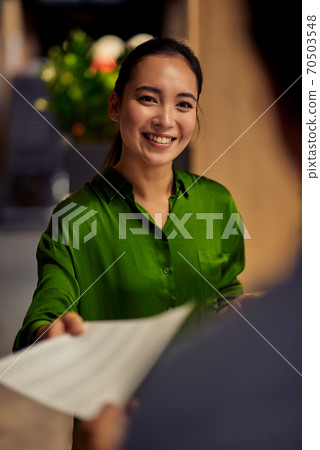 Vertical shot of a young happy asian female office worker giving some documents to her colleague and smiling while standing together in the modern office Vertical shot of a young happy asian female office worker giving some documents to her colleague and smiling while standing together in the modern office 70503548