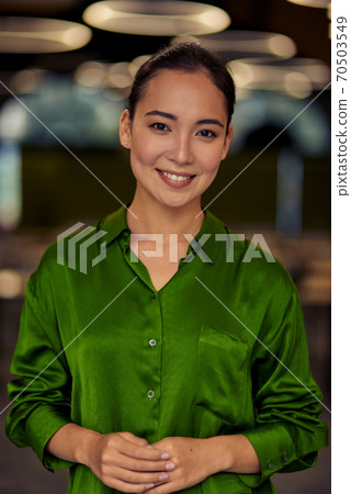 Vertical shot of a young happy asian business woman in green silk shirt looking at camera and smiling while posing in the office Vertical shot of a young happy asian business woman in green silk shirt looking at camera and smiling while posing in the office 70503549