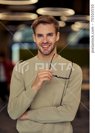 Vertical shot of a young happy caucasian businessman holding his eyeglasses, looking at camera and smiling while posing in the office Vertical shot of a young happy caucasian businessman holding his eyeglasses, looking at camera and smiling while posing in the office 70503550