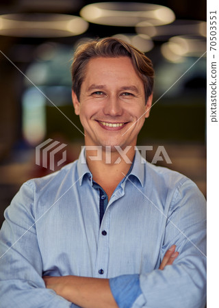 Vertical shot of a mature happy caucasian businessman in blue shirt keeping arms crossed, looking at camera and smiling while posing in the office Vertical shot of a mature happy caucasian businessman in blue shirt keeping arms crossed, looking at camera and smiling while posing in the office 70503551