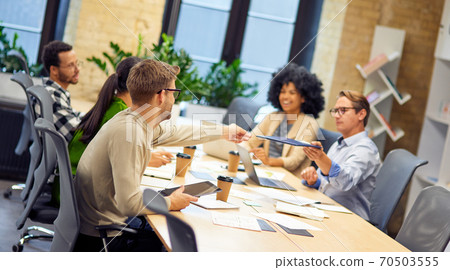 Caucasian man giving a folder with documents to his young colleague while having a meeting with project team in the modern office Caucasian man giving a folder with documents to his young colleague while having a meeting with project team in the modern office 70503555