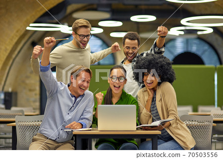 Group of young happy multiracial business people sitting at desk in the modern office or coworking space, looking at laptop screen and celebrating success Group of young happy multiracial business people sitting at desk in the modern office or coworking space, looking at laptop screen and celebrating success 70503556