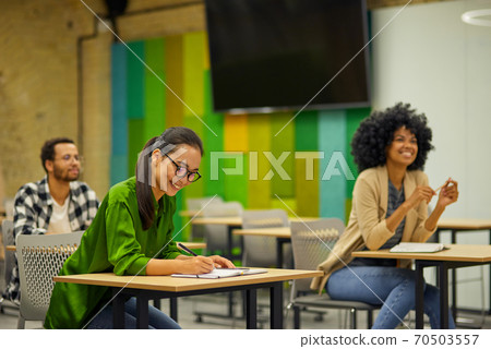 Group of young multiracial people sitting at desks in the modern office and listening to coach or speaker during corporate team building training or business seminar Group of young multiracial people sitting at desks in the modern office and listening to coach or speaker during corporate team building training or business seminar 70503557