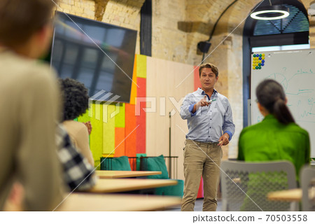 Team Building Training. Confident male business coach or speaker giving a speech while standing against multiracial group of people sitting at desks in the modern office 70503558