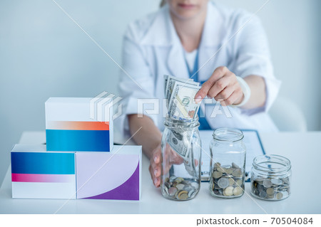 Woman in white coat sitting at the table and putting coins into the jar Woman in white coat sitting at the table and putting coins into the jar 70504084