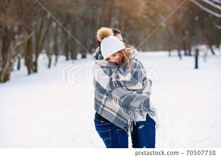 Beautiful couple hugging each other and covered with checkered plaid while walking in snowy winter park. 70507738