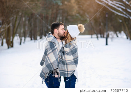 Beautiful couple hugging each other and covered with checkered plaid. Handsome man kisses his pretty girlfriend in winter park. Beautiful couple hugging each other and covered with checkered plaid. Handsome man kisses his pretty girlfriend in winter park. 70507740