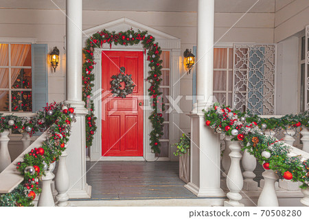 Christmas porch decoration idea. House entrance with red door decorated for holidays. Red and green wreath garland of fir tree branches and lights on railing. Christmas eve at home Christmas porch decoration idea. House entrance with red door decorated for holidays. Red and green wreath garland of fir tree branches and lights on railing. Christmas eve at home 70508280