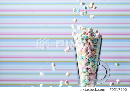 Marshmallows fall in mug. Small colorful marshmallows in glass mug on a striped background. Marshmallows fall in mug. Small colorful marshmallows in glass mug on a striped background. 70517390