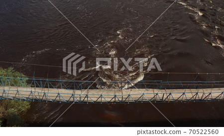 Suspended footbridge across the river. Suspended wooden pedestrian bridge across the river. bridge in the forest. green trees, foliage of trees, summer days.old hanging footbridge across a small river 70522560