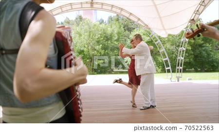 Accordion and violin playing in the park. A woman in a red dress and a man in a white suit are dancing tango. An elderly couple dancing tango in the park. 70522563