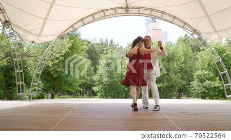 Accordion and violin playing in the park. A woman in a red dress and a man in a white suit are dancing tango. An elderly couple dancing tango in the park. 70522564