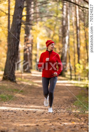 Adult caucasian slim woman in red sportswear and a cap jogging in the forest in autumn outdoor, vertical photo, selective focus 70524827