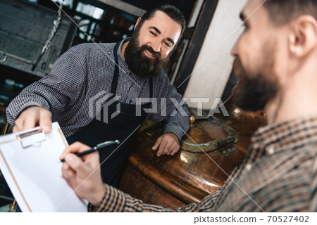 Contented bearded brewer in apron gives documents. Contented bearded brewer in apron gives documents. 70527402