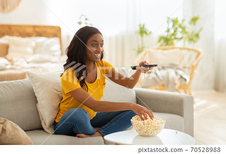 Young African American woman sitting on couch, eating popcorn and watching interesting show on television, indoors 70528988