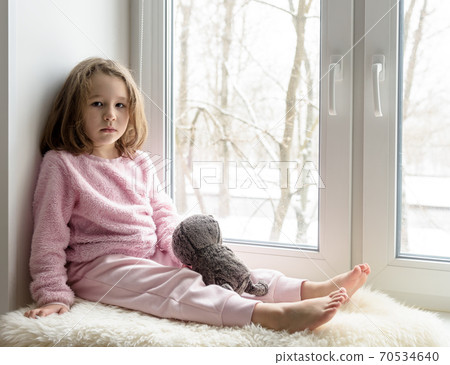 Kid sits on windowsill at home and looks at camera, portrait of pretty little girl on fur rug on room sill in winter 70534640