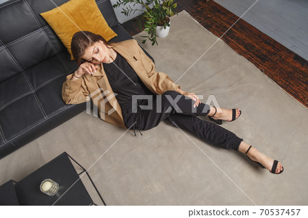 Stylishly dressed young caucasian woman in beige blazer and black pants sitting on the floor at sofa. Interior fashion portrait Stylishly dressed young caucasian woman in beige blazer and black pants sitting on the floor at sofa. Interior fashion portrait 70537457