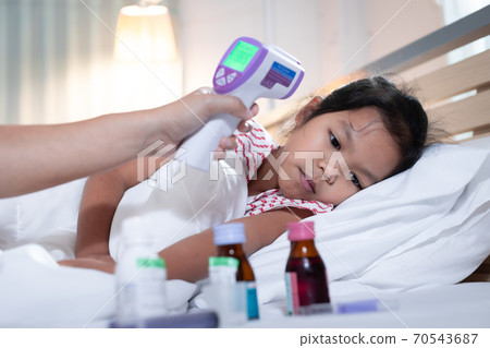 Sick asian child girl is sleeping on bed and her mother is checking up and measuring her body temperature with contactless thermometer in the bedroom at home. 70543687