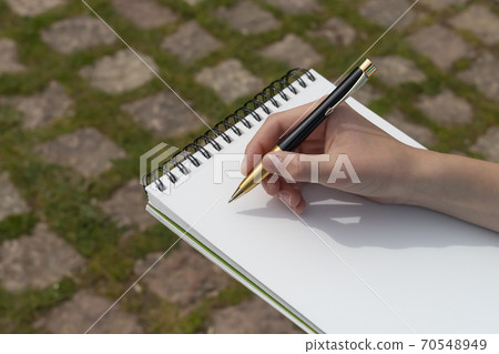 Boy's hand with a yellow-black pen over an open notepad on the background of paving stones and green grass in the park. Clean white sheet. Going to write or draw a picture. The concept of life plannin Boy's hand with a yellow-black pen over an open notepad on the background of paving stones and green grass in the park. Clean white sheet. Going to write or draw a picture. The concept of life plannin 70548949