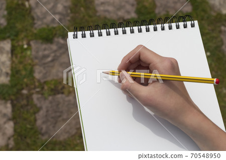 The boy's hand with a yellow-black pencil over an open notepad on the background of paving stones in the park. Clean white sheet. Going to write or draw a picture. The concept of life planning. The boy's hand with a yellow-black pencil over an open notepad on the background of paving stones in the park. Clean white sheet. Going to write or draw a picture. The concept of life planning. 70548950