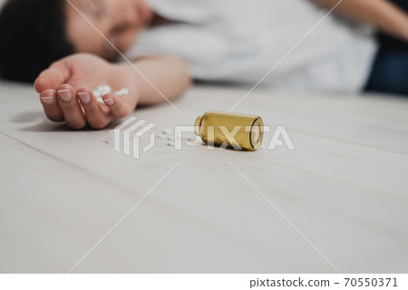In the foreground, a hand with a handful of pills in the palm lies lifeless on the floor. Nearby is a bottle of pills. Against the background of the body of a young woman. The consequences of 70550371