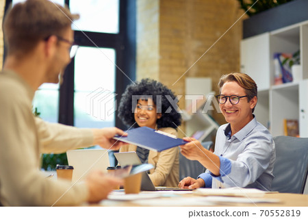 Cheerful man giving a folder with documents to his colleague and smiling while working together in the modern office 70552819