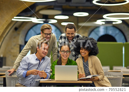 Group of young multiracial business people sitting at desk in the modern coworking space, looking at laptop screen and smiling, working together on a new project 70552821