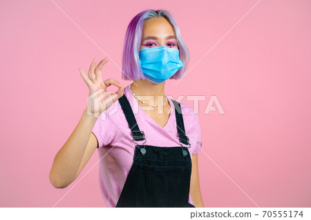 Cute woman with violet dyed hairstyle and medical mask showing okay sign over pink background Cute woman with violet dyed hairstyle and medical mask showing okay sign over pink background 70555174