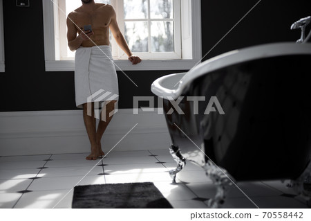 Handsome man wrapped in towel holding on mobile phone in bath Handsome man wrapped in towel holding on mobile phone in bath 70558442