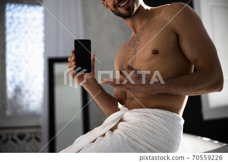 Cropped sjot of man wrapped in towel showing mobile phone on bathroom 70559226